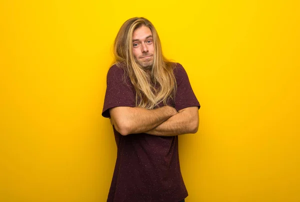 Blond man with long hair over yellow wall making doubts gesture while lifting the shoulders