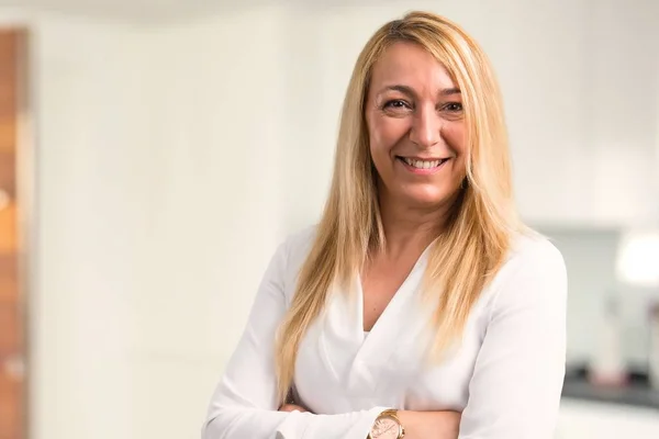 Middle age blonde woman with white shirt keeping the arms crossed in frontal position. Confident expression at home