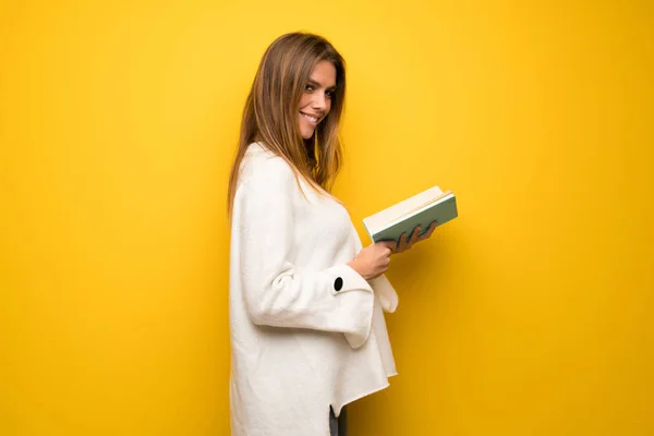 Blonde woman over yellow wall holding a book and enjoying reading