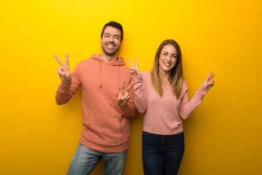 Grup gülümseyen ve iki elini zafer işaretiyle gösterilen sarı zemin üzerine iki insan