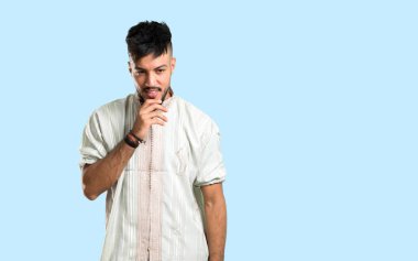 Tipik Arap kıyafetleri giyen Arap genç adam dikilip mavi arka plana bakıyor.