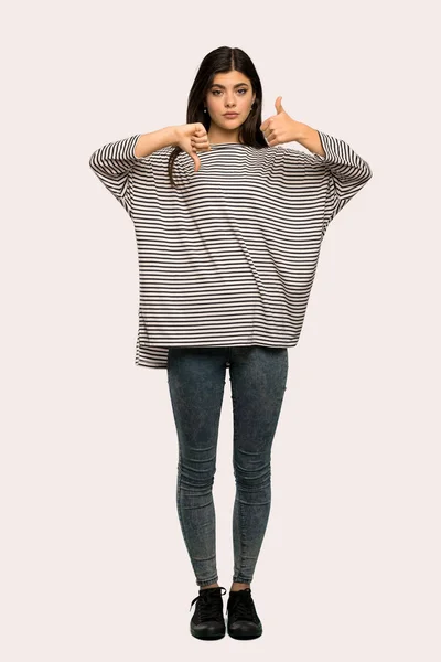 A full-length shot of a Teenager girl with striped shirt making good-bad sign. Undecided between yes or not over isolated background