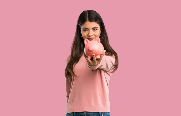 Teenager girl with pink shirt holding a piggybank on isolated pink background