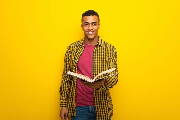 Young afro american man on yellow background holding a book and giving it to someone