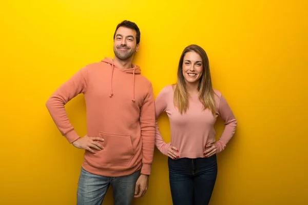 Group of two people on yellow background posing with arms at hip and laughing looking to the front