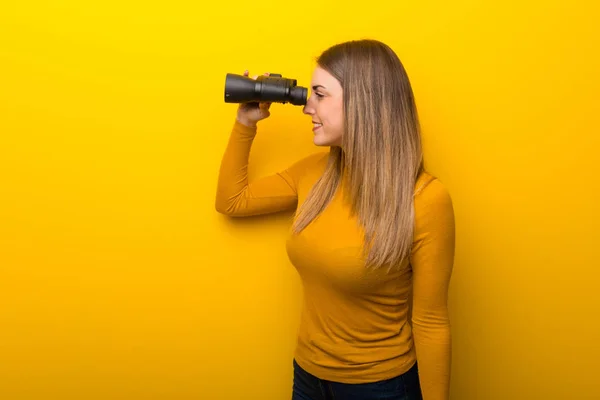 黄色の背景との距離で双眼鏡で見て若い女性 — ストック写真