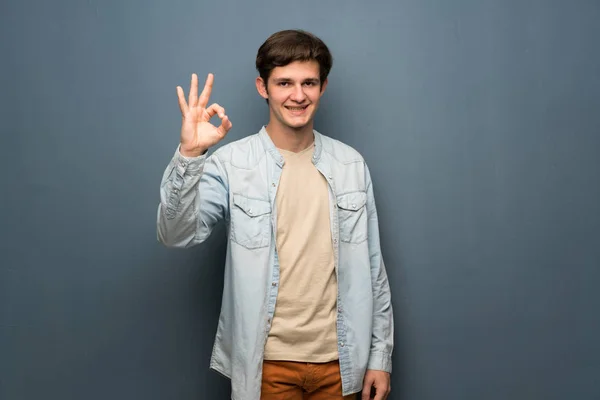 Teenager man with jean jacket over grey wall showing ok sign with fingers