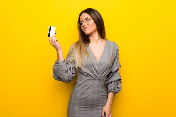 Młoda Kobieta Okularami Żółte Ściany Trzymając Kartę Kredytową Myślenia — Zdjęcie stockowe