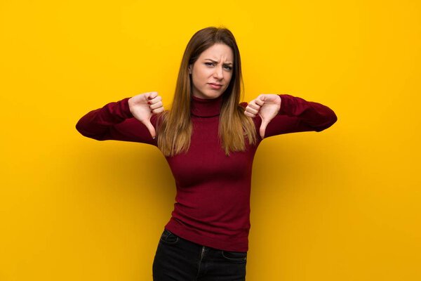 Woman with turtleneck over yellow wall showing thumb down