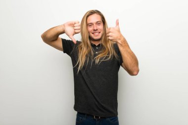 İyi-kötü işaret yapma beyaz duvarın üzerinden uzun saçlı sarışın adam. Arasında kararsız Evet ya da değil