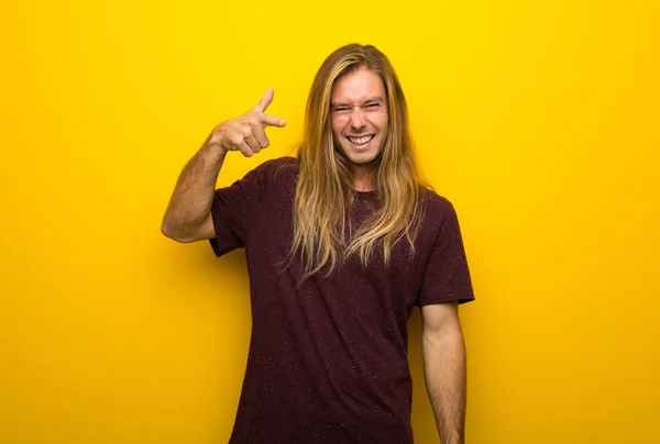 Blond man with long hair over yellow wall frustrated by a bad situation and pointing to the front