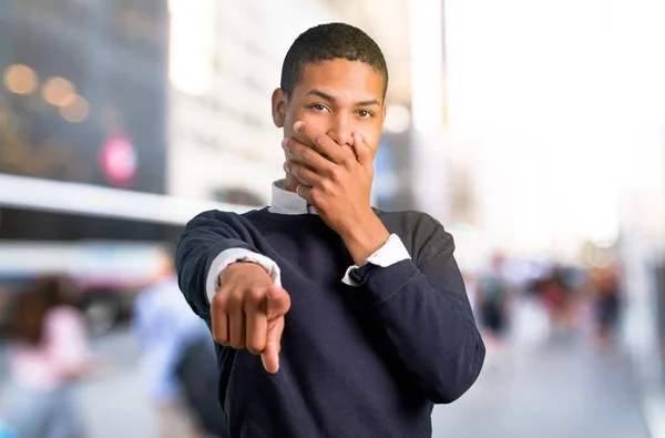Young african american man pointing with finger at someone and laughing a lot in the middle of the city