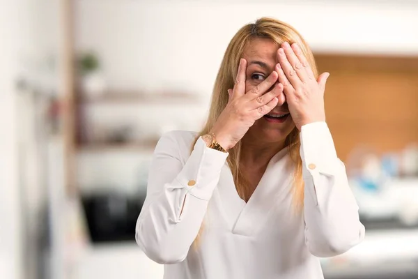 Middle age blonde woman with white shirt covering eyes by hands and looking through the fingers at home