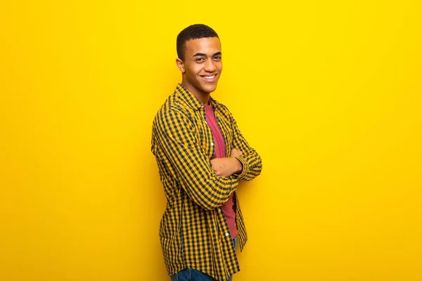 Young afro american man on yellow background keeping the arms crossed in lateral position while smiling