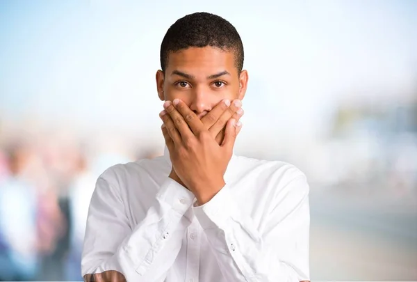 Young african american man covering mouth with both hands for saying something inappropriate. Can not speak on unfocused outdoor background