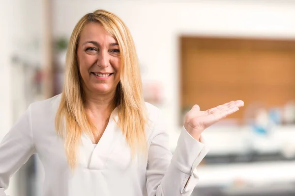 Middle age blonde woman with white shirt holding copyspace imaginary on the palm to insert an ad at home