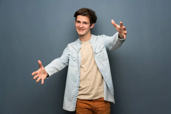 Teenager man with jean jacket over grey wall presenting and inviting to come with hand