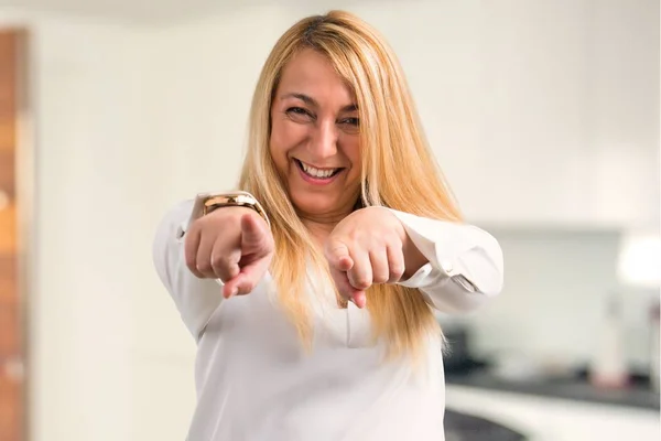 Middle age blonde woman with white shirt points finger at you while smiling at home