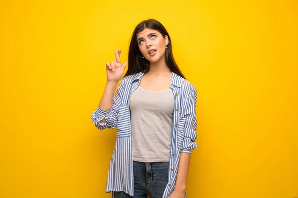 Teenager girl over yellow wall with fingers crossing and wishing the best