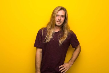 Silah, kalça ile poz ve gülümseyen sarı duvarın üzerinden uzun saçlı sarışın adam