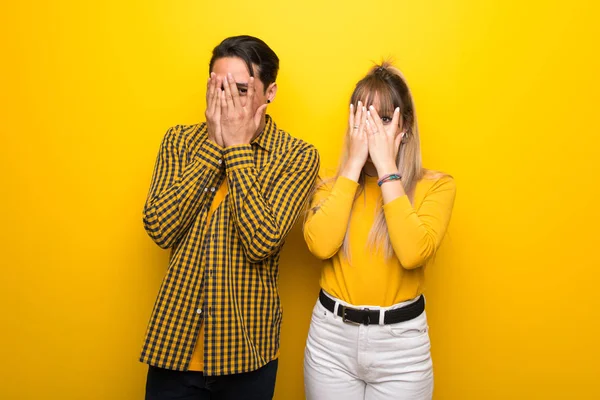 Canlı sarı arka plan üzerinde genç çift elleri ile gözleri kaplayan ve parmaklardan bakarak