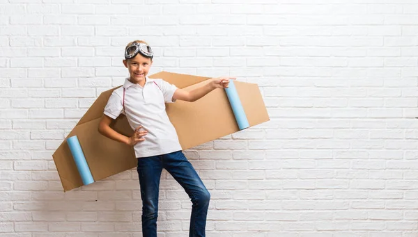 Boy playing with cardboard airplane wings on his back pointing finger to the side and presenting a product