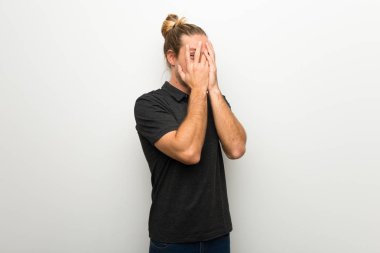 Gözleri eller tarafından kapsayan ve parmaklarının arasından seyir beyaz duvarın üzerinden uzun saçlı sarışın adam