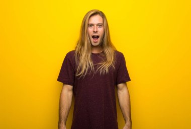 Blond man with long hair over yellow wall with surprise and shocked facial expression clipart