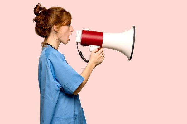Young redhead nurse shouting through a megaphone over pink background