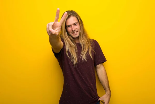 笑顔と勝利のサインを示す黄色の壁に長い髪の金髪の男 — ストック写真