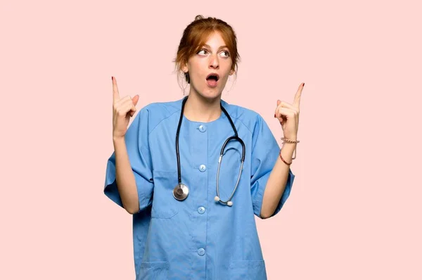 Joven Pelirroja Enfermera Sorprendida Apuntando Hacia Arriba Sobre Fondo Rosa — Foto de Stock