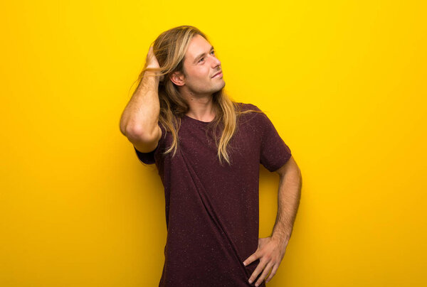 Блондин с длинными волосами над жёлтой стеной думает об идее, пока чешет голову.
