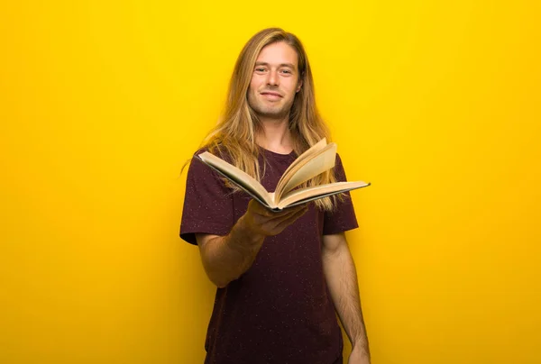本を持って 誰かにそれを与える黄色の壁に長い髪の金髪の男 — ストック写真