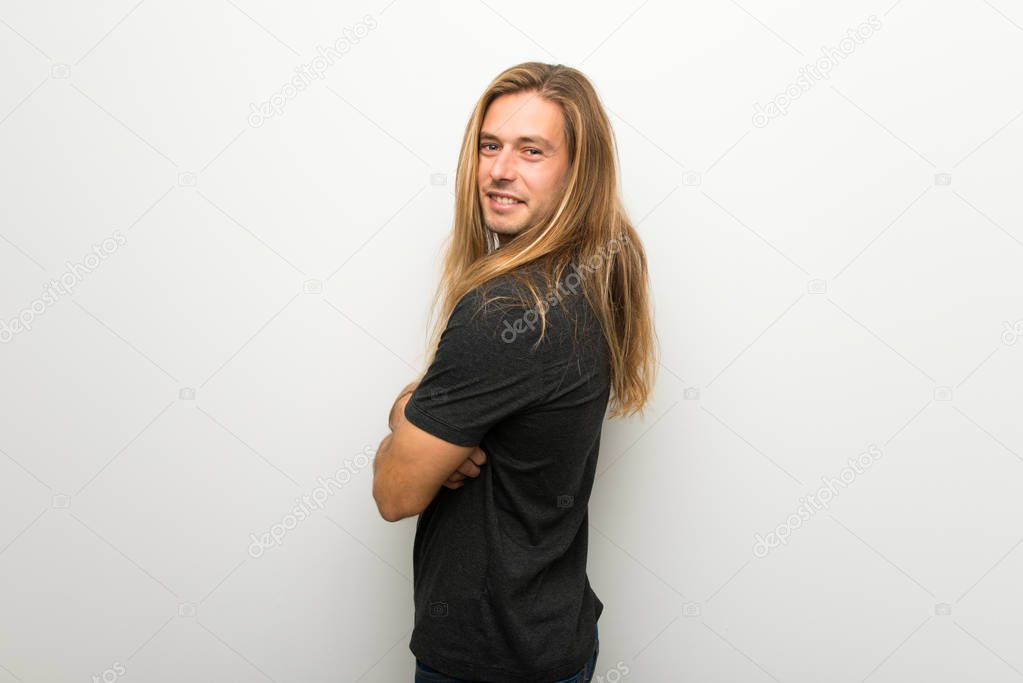Blond man with long hair over white wall looking over the shoulder with a smile