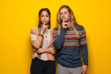 Hippi çift bir işaret parmağını ağzına koyarak sessizlik jest gösteren sarı arka plan üzerinde