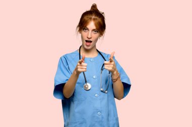 Öne doğru işaret ve pembe arka plan üzerinde gülümseyen genç redhead hemşire