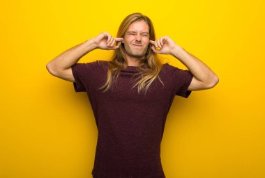 Her iki kulak elleriyle kapsayan sarı duvarın üzerinden uzun saçlı sarışın adam