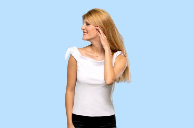 Sarışın genç kadın için bir şey kulak izole mavi zemin üzerine elini koyarak dinleme