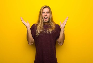 Sürpriz ve şok yüz ifadesi ile sarı duvarın üzerinden uzun saçlı sarışın adam