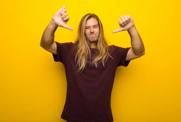 両手でダウン親指を示す黄色の壁に長い髪の金髪の男 — ストック写真