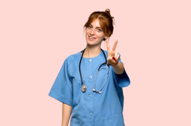 Gülümseyen ve zafer işareti pembe arka plan üzerinde gösterilen genç redhead hemşire