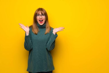 Sürpriz ve şok yüz ifadesi ile sarı duvarın üzerinden gözlüklü kadın