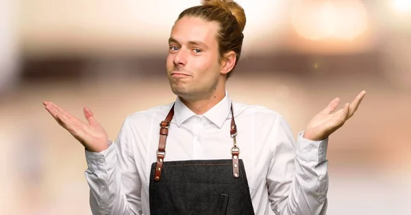 Barber man in an apron having doubts while raising hands and shoulders in a barber shop