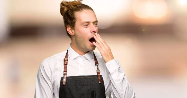 Barber man in an apron yawning and covering wide open mouth with hand in a barber shop