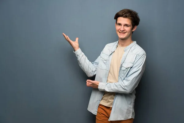 Teenager man with jean jacket over grey wall extending hands to the side for inviting to come