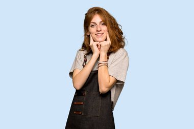 Mavi arka plan üzerinde mutlu ve keyifli bir ifadeyle gülümseyen önlük ile genç Kızıl saçlı kadın