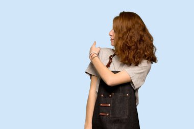 Mavi arka plan üzerinde geri işaret parmağı ile işaret önlük ile genç Kızıl saçlı kadın