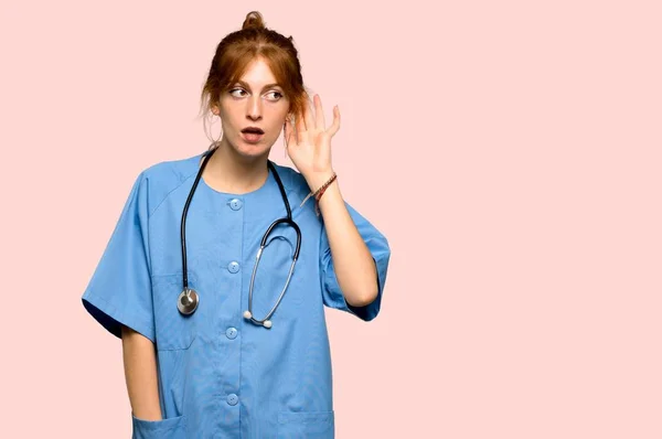 Young Redhead Nurse Listening Something Putting Hand Ear Pink Background — Stock Photo, Image