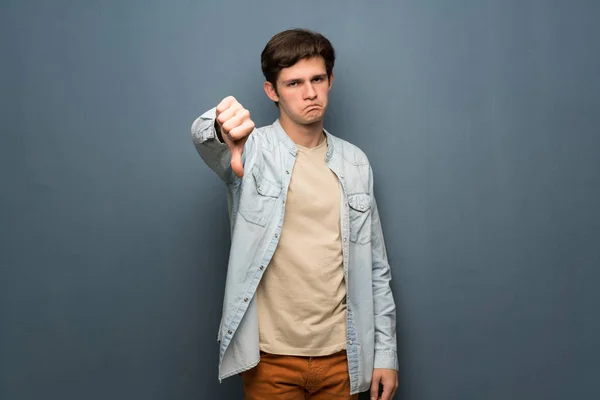 Teenager man with jean jacket over grey wall showing thumb down with negative expression
