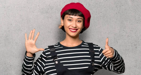 Young woman with beret counting six with fingers over textured wall